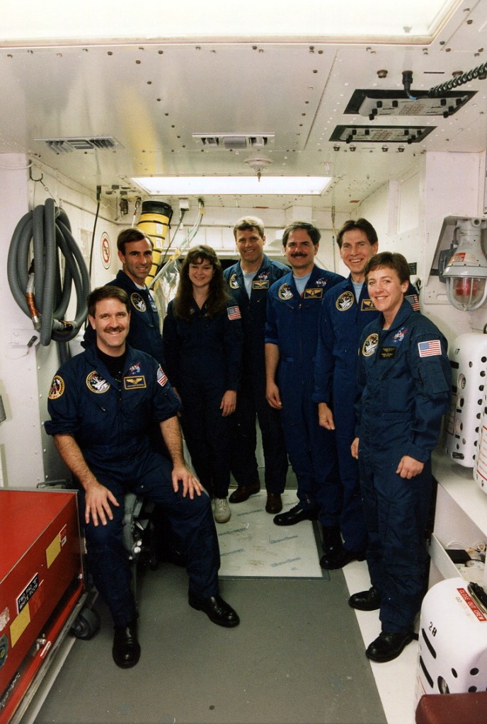 Group photo of seven astronauts wearing blue flight suits in the White Room at the launch pad.