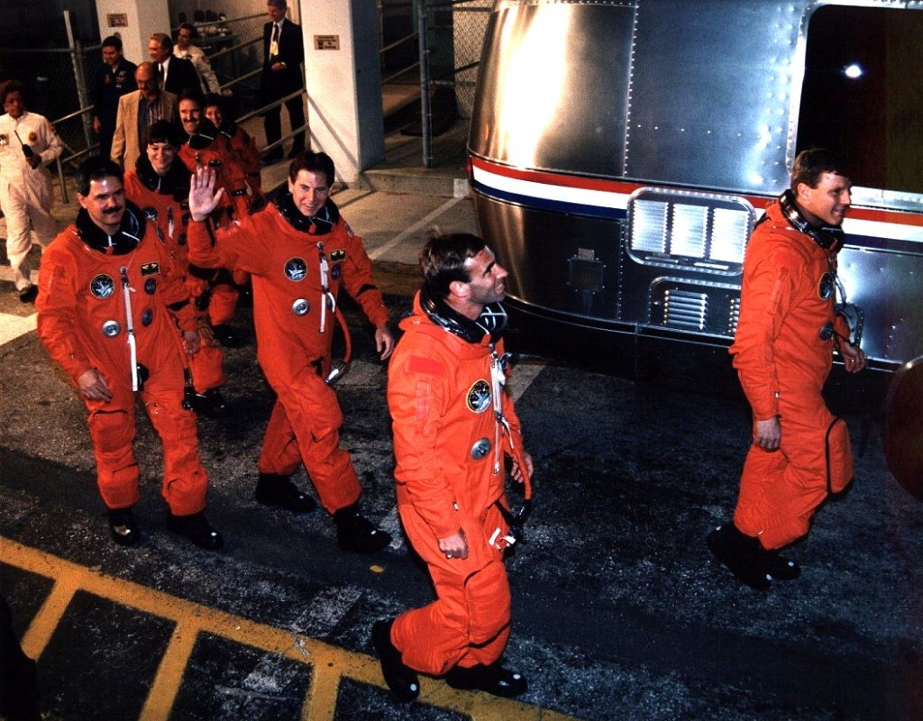 A group of seven astronauts wearing orange spacesuits without helmets walk out of a building to board a silver van.