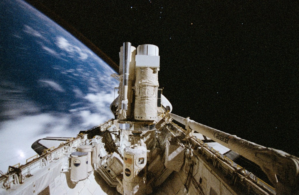 Image of a space shuttle payload bay with several telescopes pointing out of the bay toward a black sky, with a portion of the Earth visible.