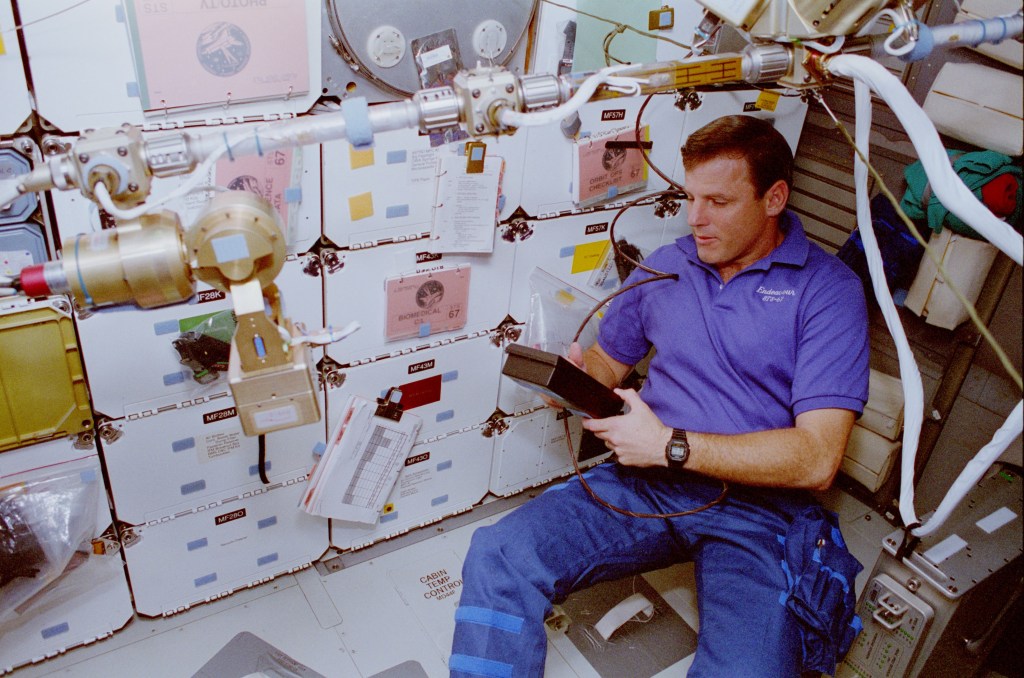 Image of a male astronaut wearing a blue polo shirt and working on an experiment in the shuttle middeck.